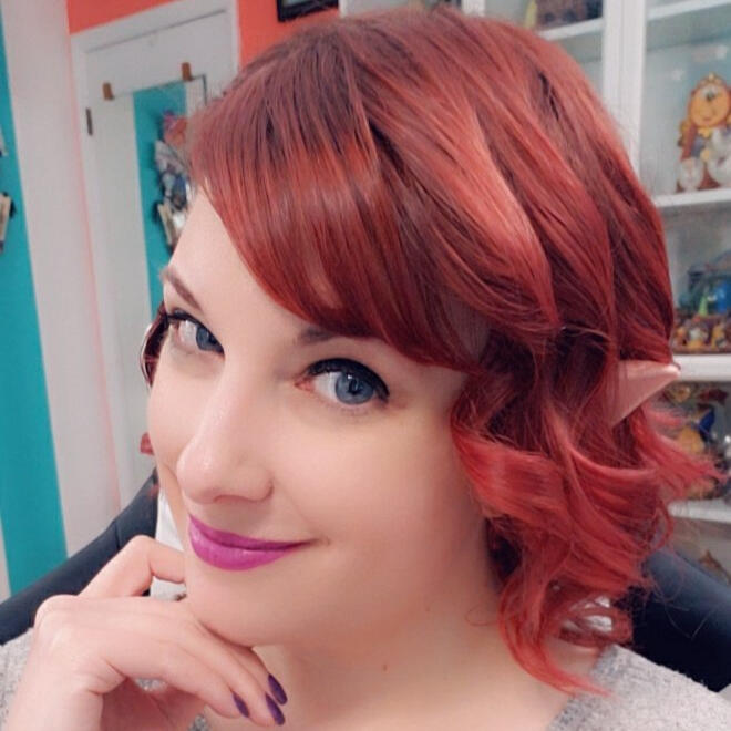 A white woman with red hair smiles at the camera. Her hair is curled and the hint of fake elf ears are poking out through the hair. She is wearing a flower crown of white and pink flowers.