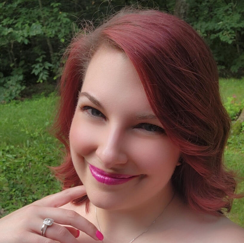 A white woman with red hair smiles at the camera. Her hair is curled and the hint of fake elf ears are poking out through the hair. She is wearing a flower crown of white and pink flowers.