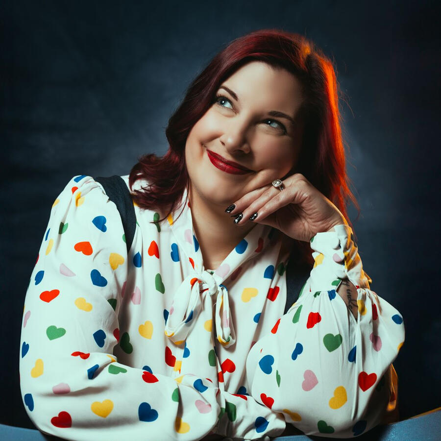 A white woman with red hair smiles at the camera. Her hair is curled and the hint of fake elf ears are poking out through the hair. She is wearing bright pink lipstick.